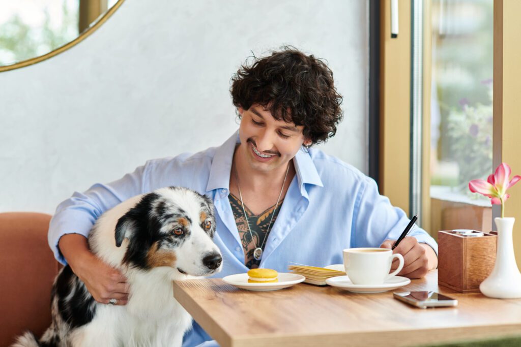 Man with curly hair and tattoos writing in a notebook at a cafe with a dog by his side and a plate with a macaron and a cup of coffee on the table