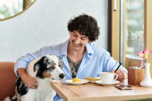 Man with curly hair and tattoos writing in a notebook at a cafe with a dog by his side and a plate with a macaron and a cup of coffee on the table
