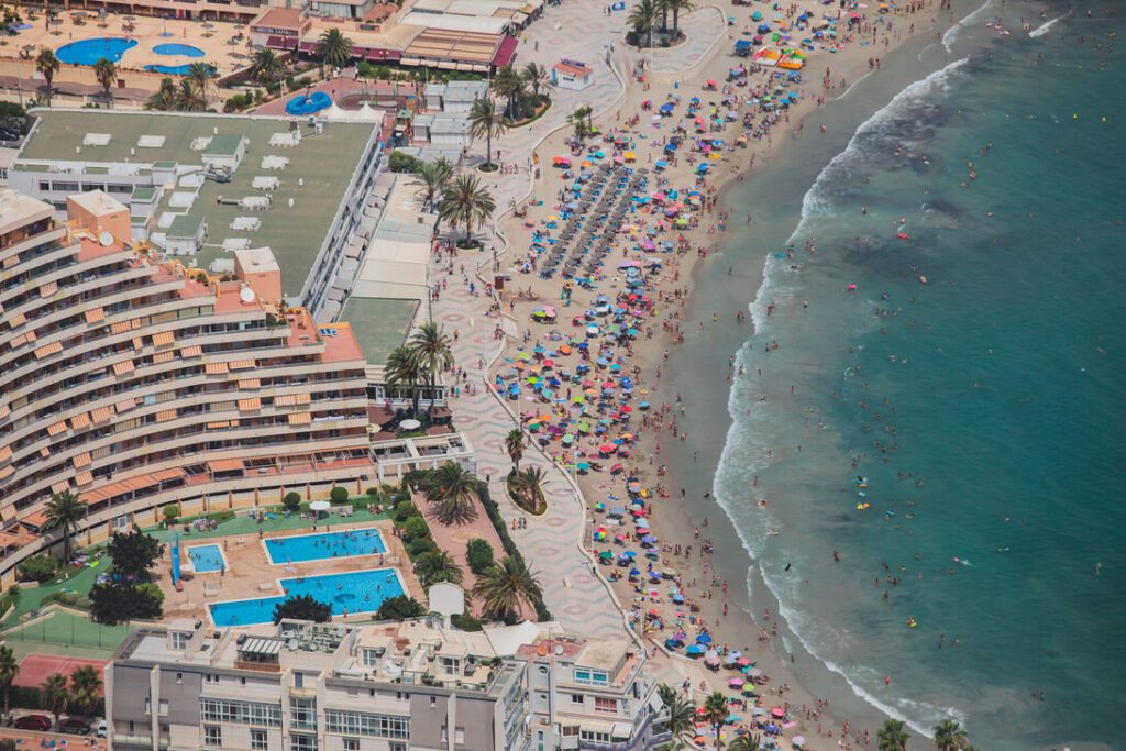 Aerial view of a crowded beach resort with a curved hotel sun loungers palm trees and a busy shoreline with colorful umbrellas and swimmers