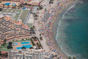 Aerial view of a crowded beach resort with a curved hotel sun loungers palm trees and a busy shoreline with colorful umbrellas and swimmers
