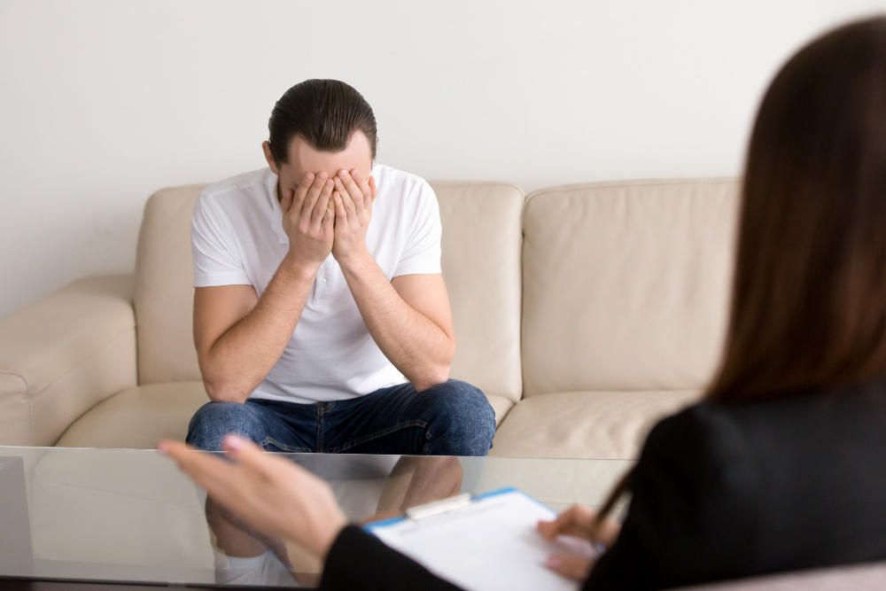 Distressed man sitting on a beige sofa hands over his face during a therapy session with a counselor nearby