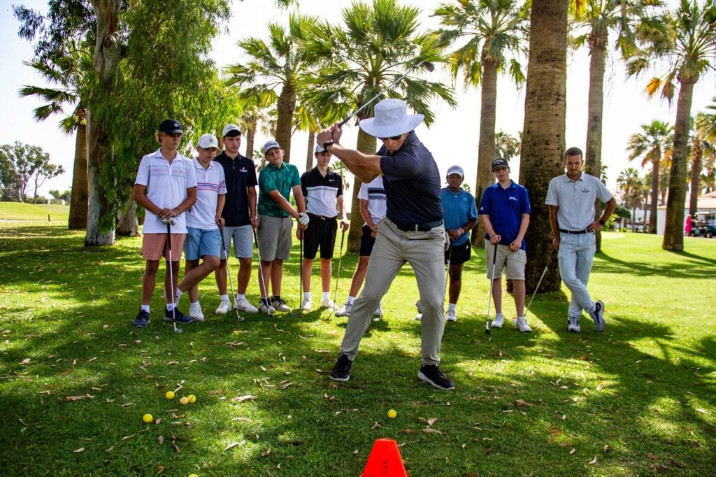 Instructor in a white sun hat demonstrates a golf swing to a group of young players on a sunny, palm-lined course. An orange cone sits on the grass in front of them.