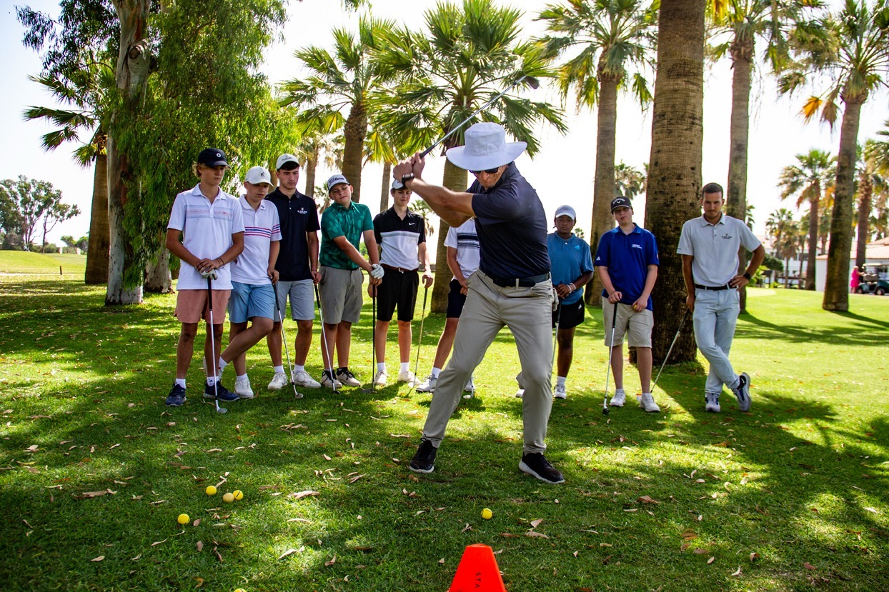 772284 Black Iron Andalucía Noticias 24h Instructor in a white sun hat demonstrates a golf swing to a group of young players on a sunny palm lined course An orange cone sits on the grass in front of them