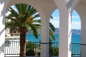 White arched colonnade framing a palm tree and a blue sea beyond with a black railing in the foreground