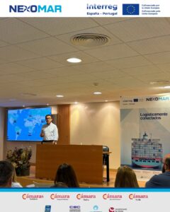 Speaker presenting at a conference stage with a world map on a screen banner reading Logísticamente conectados and logos Interreg NEXOMAR audience seated in a lecture hall