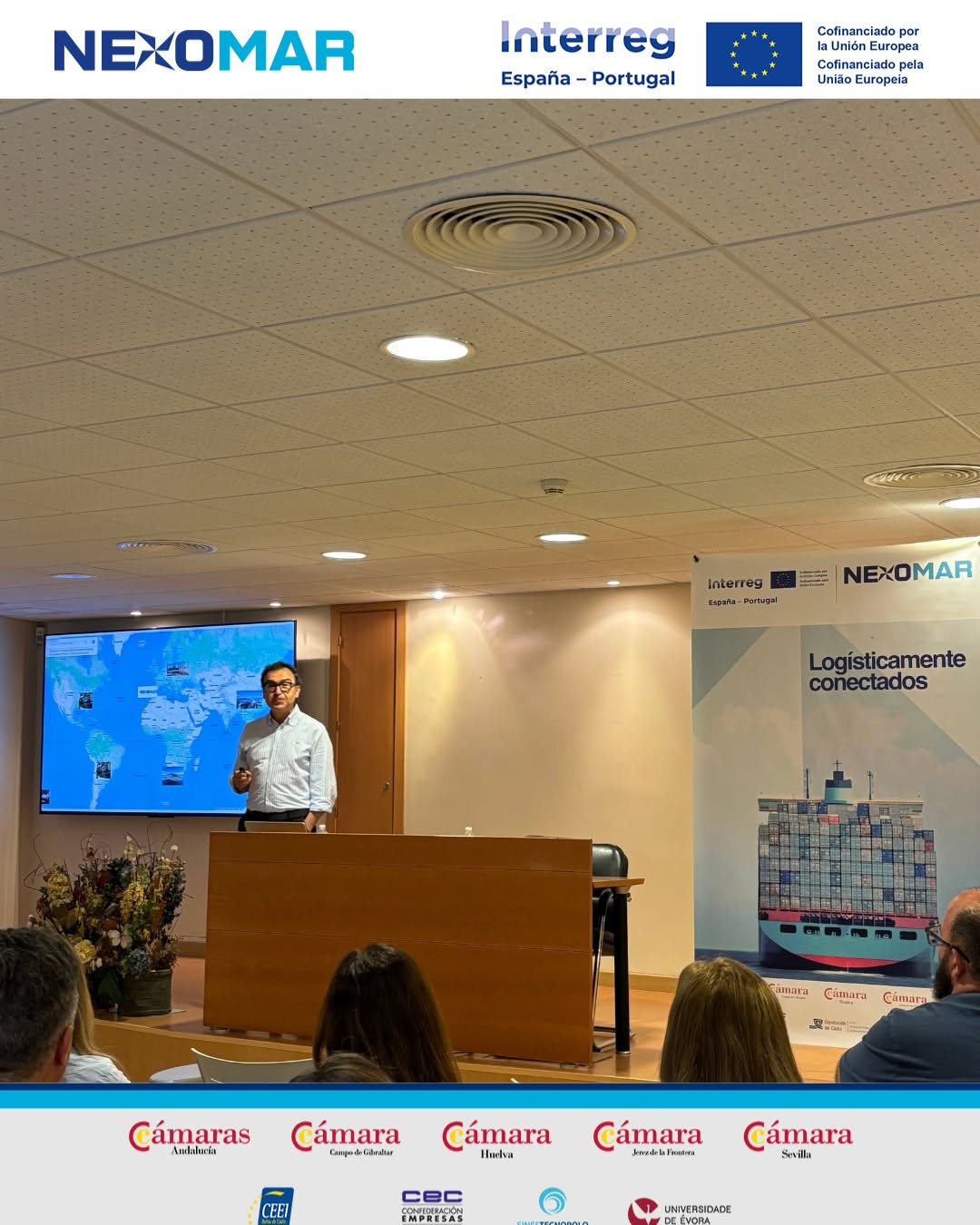 Nexomar IP Andalucía Noticias 24h Speaker presenting at a conference stage with a world map on a screen banner reading Logísticamente conectados and logos Interreg NEXOMAR audience seated in a lecture hall