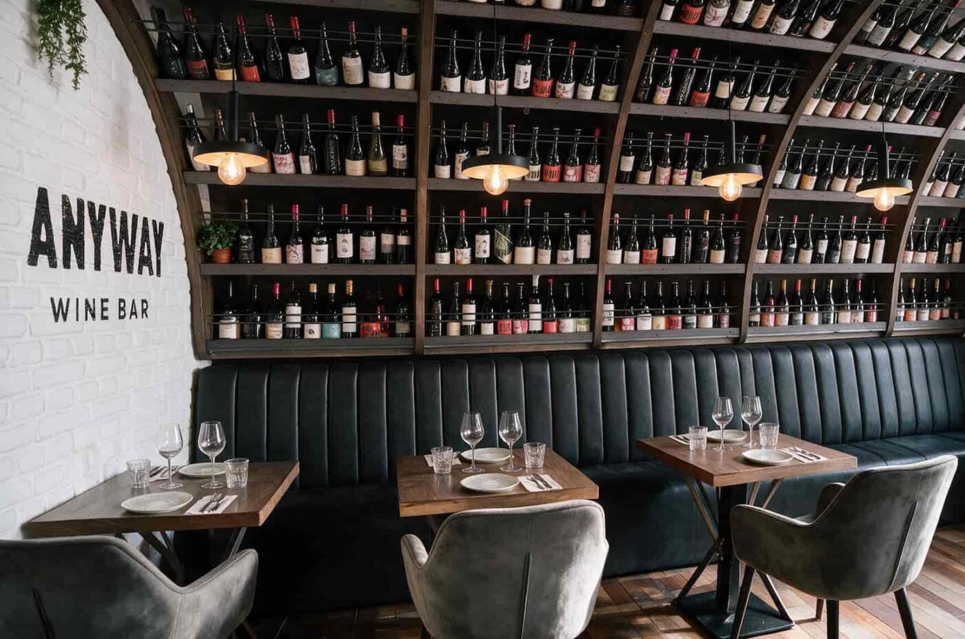 anywaywinebar IP Andalucía Noticias 24h Wine bar interior with a dark leather banquette wooden tables set with glasses and plates and wine bottles on shelves against a white brick wall
